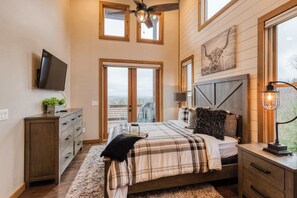 A bedroom with a bed, dresser, TV, windows, ceiling fan, and artwork.