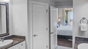 A modern bathroom with white fixtures, granite countertops, and a visible bed in the adjacent room. - Azure 223 | 3BR Coastline View | FREE Beach Service (Fort Walton Beach)