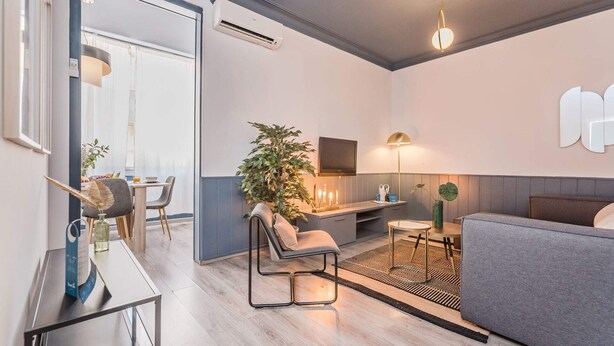 A modern living room with a grey sofa, a dining area with a table and chairs, a large potted plant, and a kitchen area with a TV and lamp. - Sweett | Rocafort Principal 1 (Barcelona)