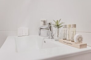 A bathroom sink with a white countertop, a glass vase with a plant, and three bottles of shampoo. - Sweett | Rocafort Principal 2 (Barcelona)