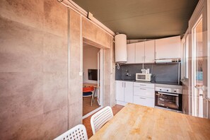 Een kleine keuken met witte kasten, een magnetron, een fornuis, en een eethoek met een houten tafel en witte stoelen.