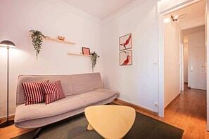 A living room with a sofa, a floor lamp, a small table, and wall-mounted shelves with plants and pictures.