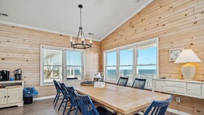Un comedor amplio con mesa de madera y sillas azules, ventanas grandes con vistas al océano y una araña.