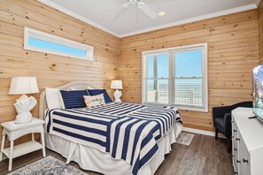 Ein Schlafzimmer mit einer holzverkleideten Wand, einem großen Fenster mit Jalousien, einem Bett mit gestreifter Bettdecke, zwei Nachttischen mit Lampen und einem Schminktisch.