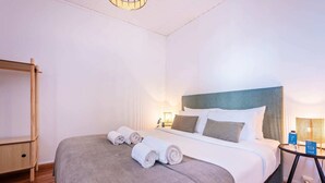 Una cama bien hecha con almohadas y toallas enrolladas, una mesita de noche con lámpara, y una estantería de madera.