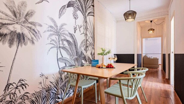 Un comedor con una mesa de madera, sillas verdes y un mural tropical en la pared.
