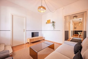 A modern living room with a flat-screen TV, a glass coffee table, and a sofa. There is a wooden cabinet and a plant on the wall.