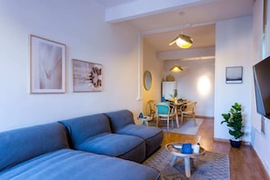 A modern living room with a grey sofa, a dining area with a table and chairs, and a TV mounted on the wall.