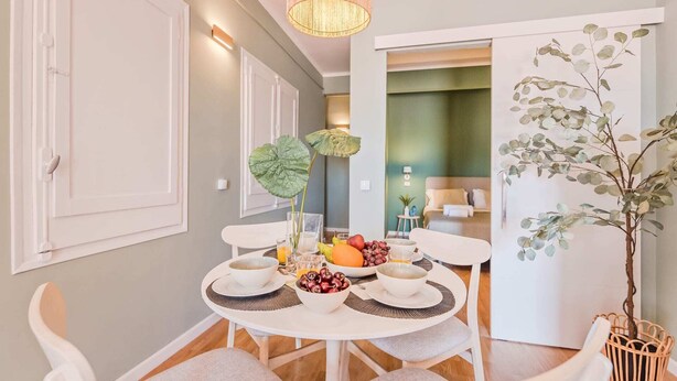 A dining area with a round table set for four, surrounded by white chairs. There is a large leafy plant on the table, and a hanging light fixture above. - Sweett | Rocafort Atic 3 (Barcelona)