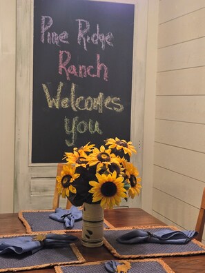 Una pizarra con 'Pine Ridge Ranch' y 'Bienvenidos' escritos, un jarrón con girasoles y una mesa con servilletas dobladas.