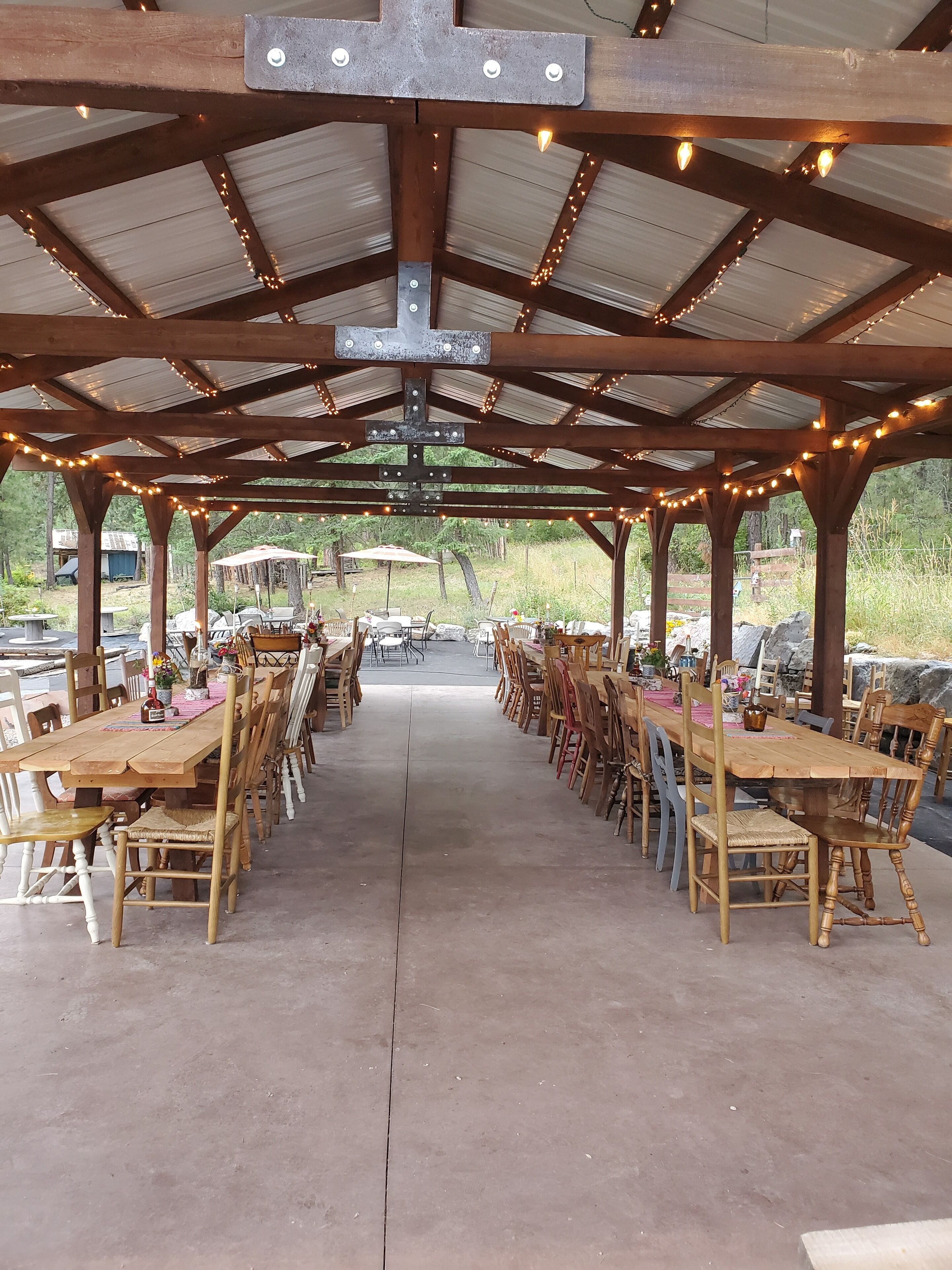 A covered outdoor dining area with wooden tables and chairs, string lights, and a view of the surrounding landscape.