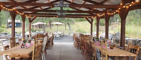 A covered outdoor dining area with wooden tables and chairs, string lights, and a view of the surrounding landscape.