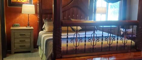 A four-poster bed with a wooden frame and metal railings, a bedside table with a lamp, a framed picture on the wall, and a window with curtains.