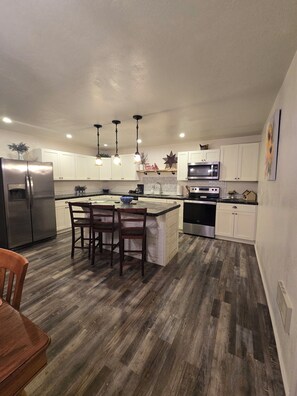 A modern kitchen with white cabinets, a stainless steel refrigerator, a microwave, and a dishwasher. There are wooden chairs around a central island with a sink.