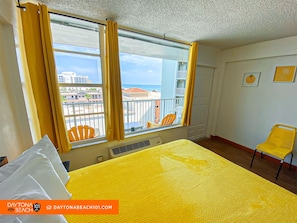 A hotel room with a large window, yellow curtains, a bed with a yellow bedspread, a chair, and a view of the city and beach. - Beachfront Studio on Daytona Beach w/ Oceanfront Pool & Free Parking (Daytona Beach)