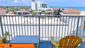 A balcony with yellow chairs and a table, overlooking a beach and a hotel with a red-tiled roof. - Beachfront Studio on Daytona Beach w/ Oceanfront Pool & Free Parking (Daytona Beach)