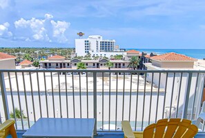 Un balcon avec des chaises jaunes et une table, donnant sur une plage et un hôtel dont le toit est recouvert de tuiles rouges.