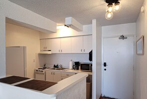 Une cuisine de taille réduite, avec des meubles blancs, une cuisinière, un four à micro-ondes et un évier. Une porte donne accès à une autre pièce.