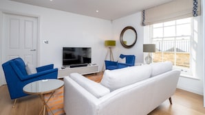 Un salon moderne avec un canapé blanc, des fauteuils bleus, un téléviseur et une grande fenêtre donnant sur un beau paysage.
