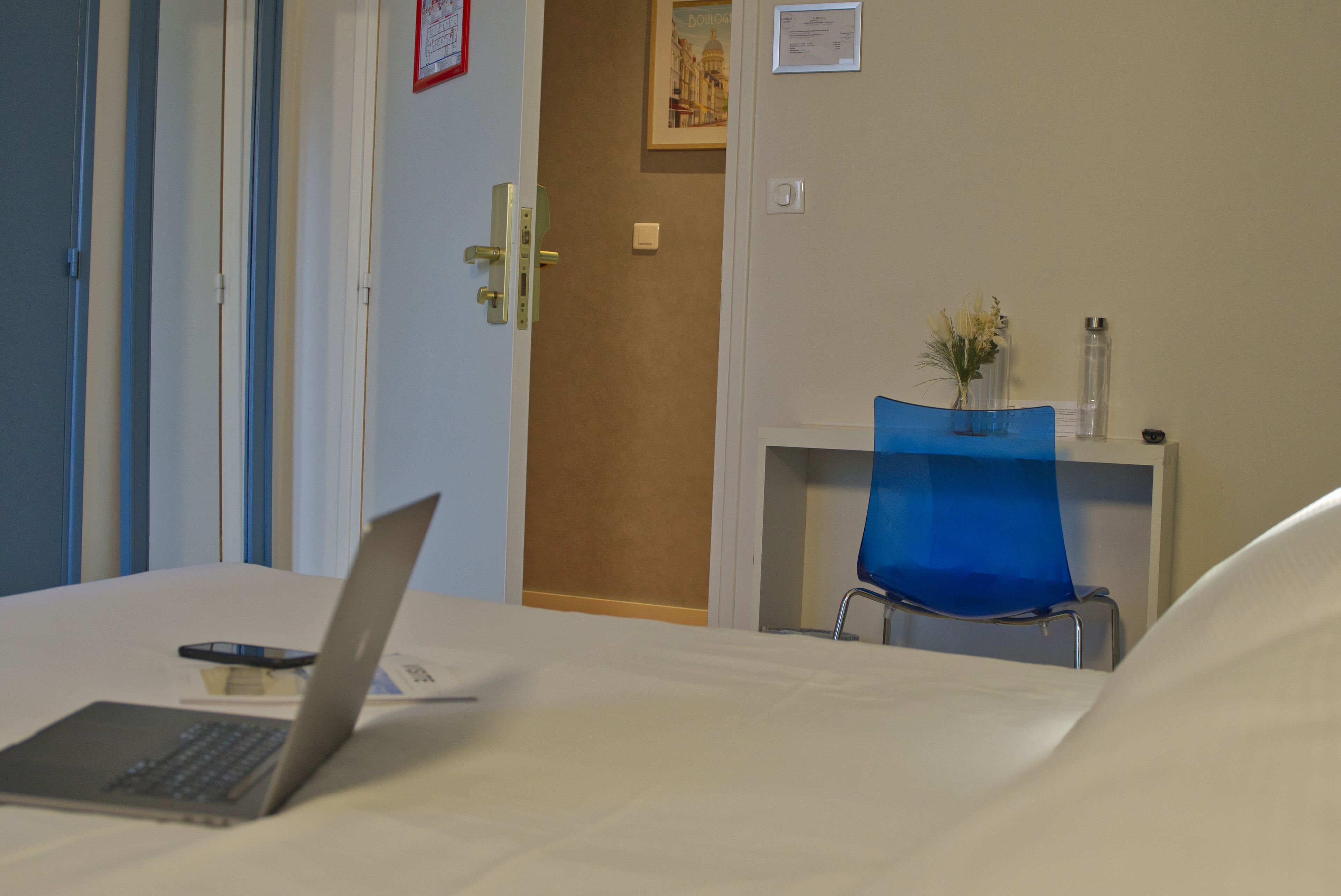 A hotel room with a bed, a laptop, a smartphone, a blue chair, a vase with flowers, and a framed picture on the wall.