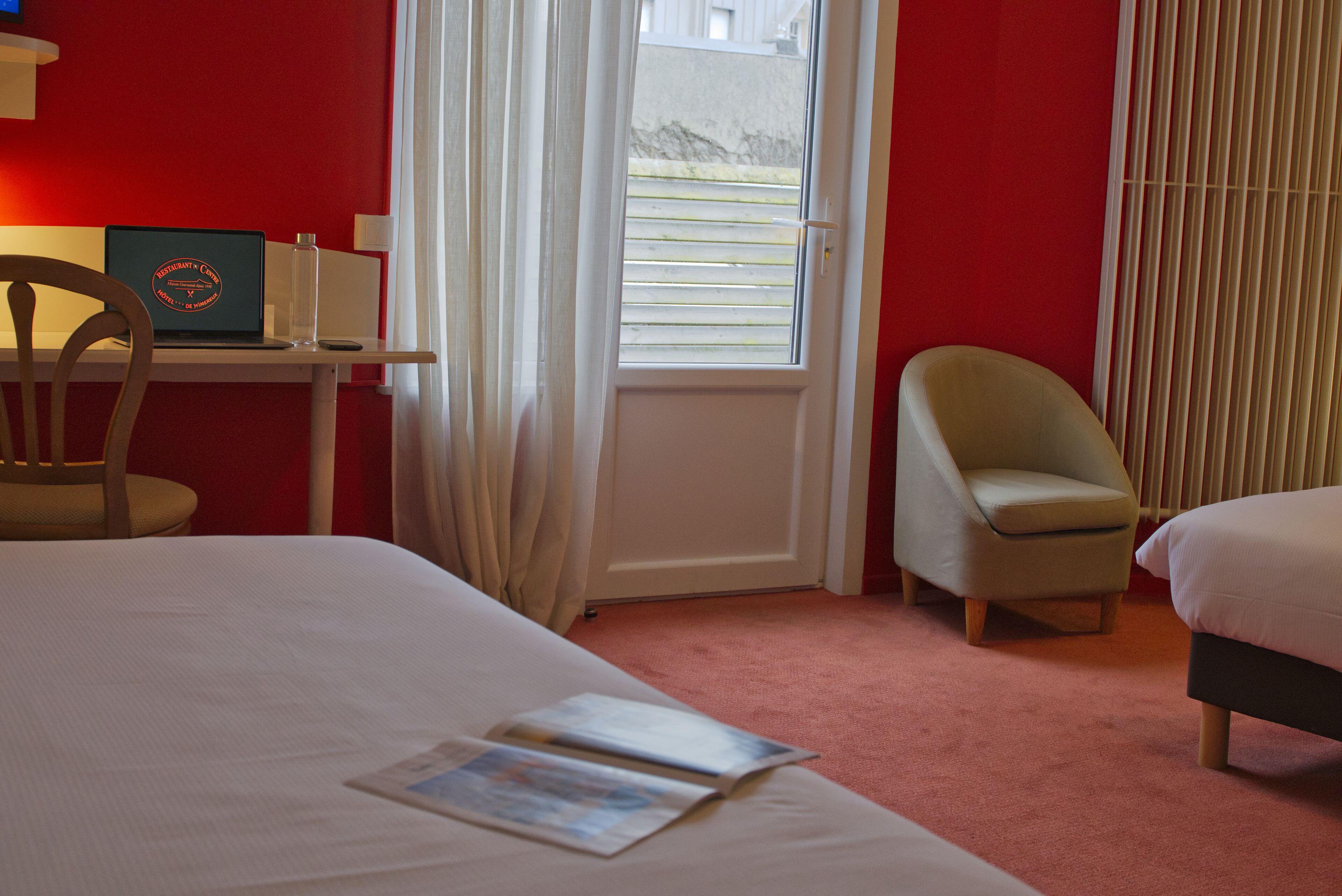 Een hotelkamer met een bed, een bureau met een laptop, een stoel en een raam met jaloezieën.