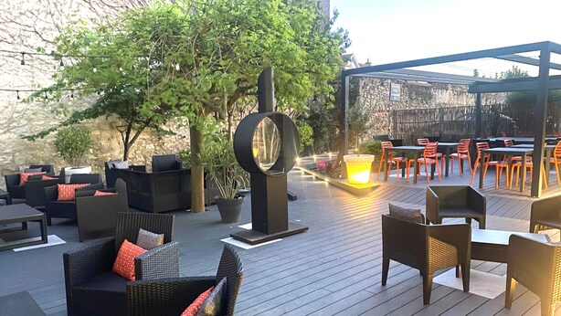 Un patio al aire libre con mobiliario de mimbre, un pilar decorativo central y un conjunto de asientos bajo una pérgola.