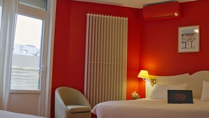 Una habitación de hotel con cama, una silla, una ventana, un radiador y una lámpara fijada a la pared.