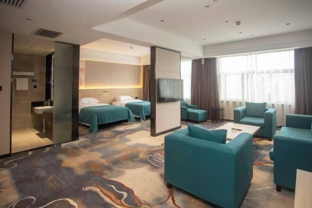 Habitación de hotel con una cama grande, dos camas más pequeñas, un sofá, un televisor y un baño con espejo y lavamanos.