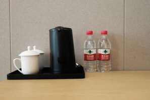Une bouilloire électrique noire, une tasse blanche et deux bouteilles d’eau avec des bouchons rouges, posées sur une table en bois devant un mur de couleur beige.