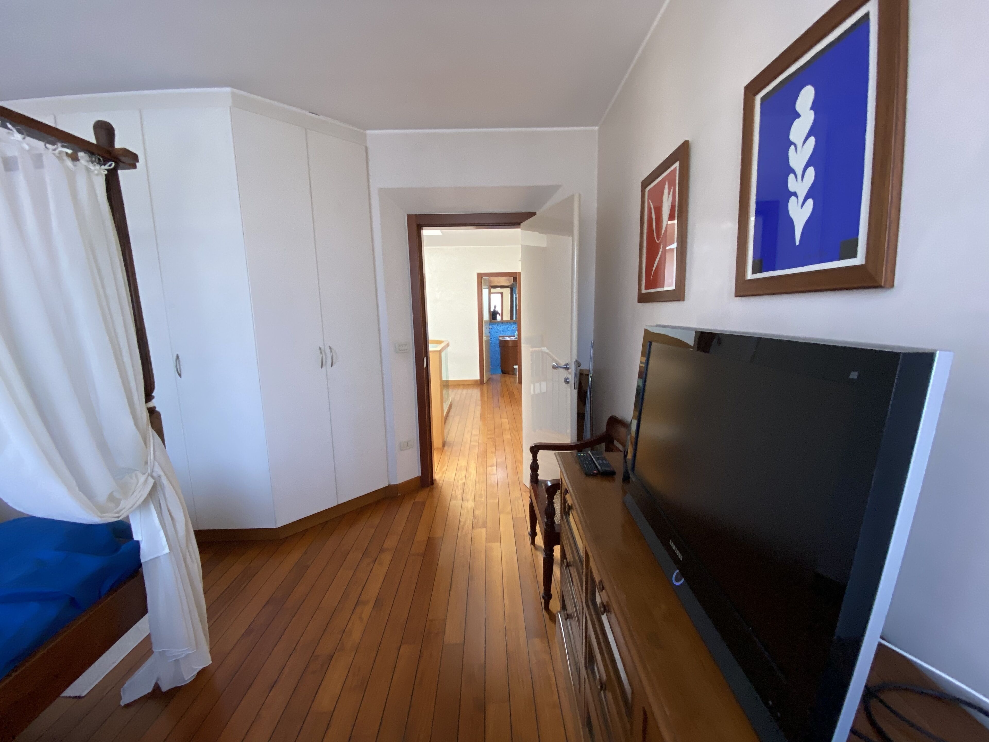 Una habitación con suelo de madera, una cama con cortinas blancas, un escritorio con una computadora y un televisor.