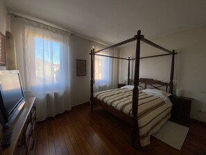 A bedroom with a four-poster bed, wooden flooring, a desk with a computer, and a window with sheer curtains.
