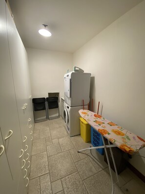 A compact kitchen with a washing machine, two trash bins, a table with a floral tablecloth, and a ceiling light.
