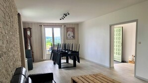 Ein Wohnzimmer mit schwarzem Ledersofa, einem Holztisch, einem Esstisch mit Stühlen, einer Uhr und Blick durch ein Fenster.