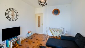 Una sala de estar con televisor de pantalla plana, un sofá, una mesa de centro, una mesa de comedor con sillas, un reloj decorativo y una lámpara colgante.
