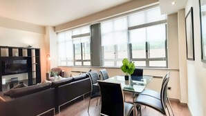 A modern living room with a sofa, a glass table, and chairs. There is a TV on a black cabinet, and a plant on the table. - 1-bed Apartment in City of London (London)