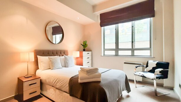 A neatly arranged bedroom with a bed, bedside tables, a mirror, a window with blinds, and a chair. - 1-bed Apartment in City of London (London)