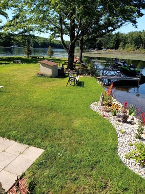 A well-maintained grassy lawn with a picnic table and a tree, a dock with a boat, and a lake in the background.