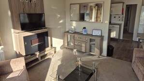 Una sala moderna con televisor de pantalla plana, chimenea, una mesa de centro de vidrio y una zona de cocina al fondo.