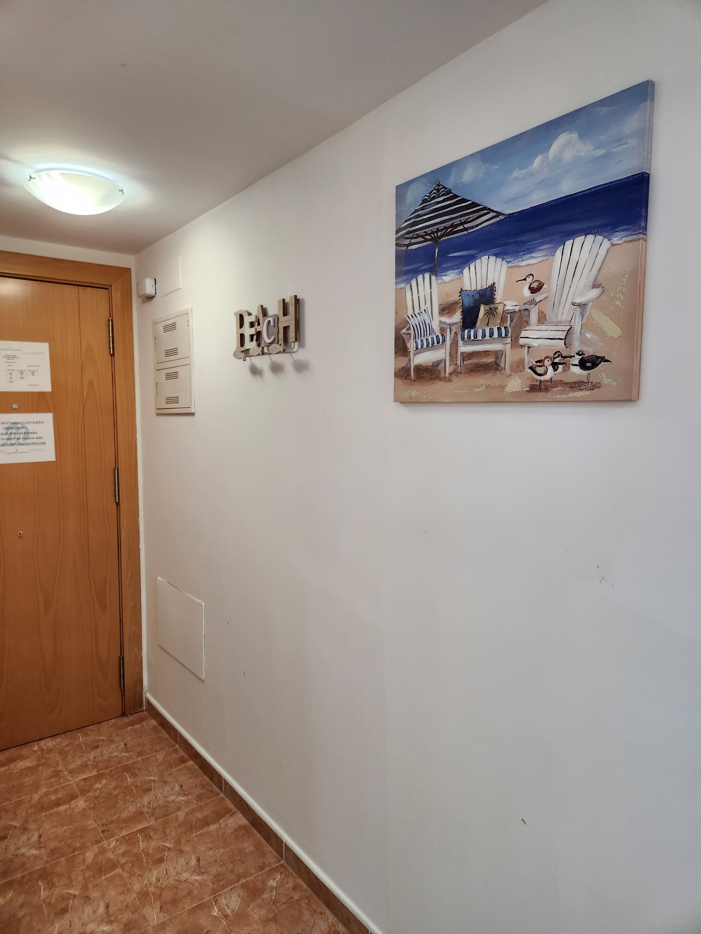 A hallway with a wooden door, a ceiling light, a wall-mounted thermostat, and a beach-themed painting.
