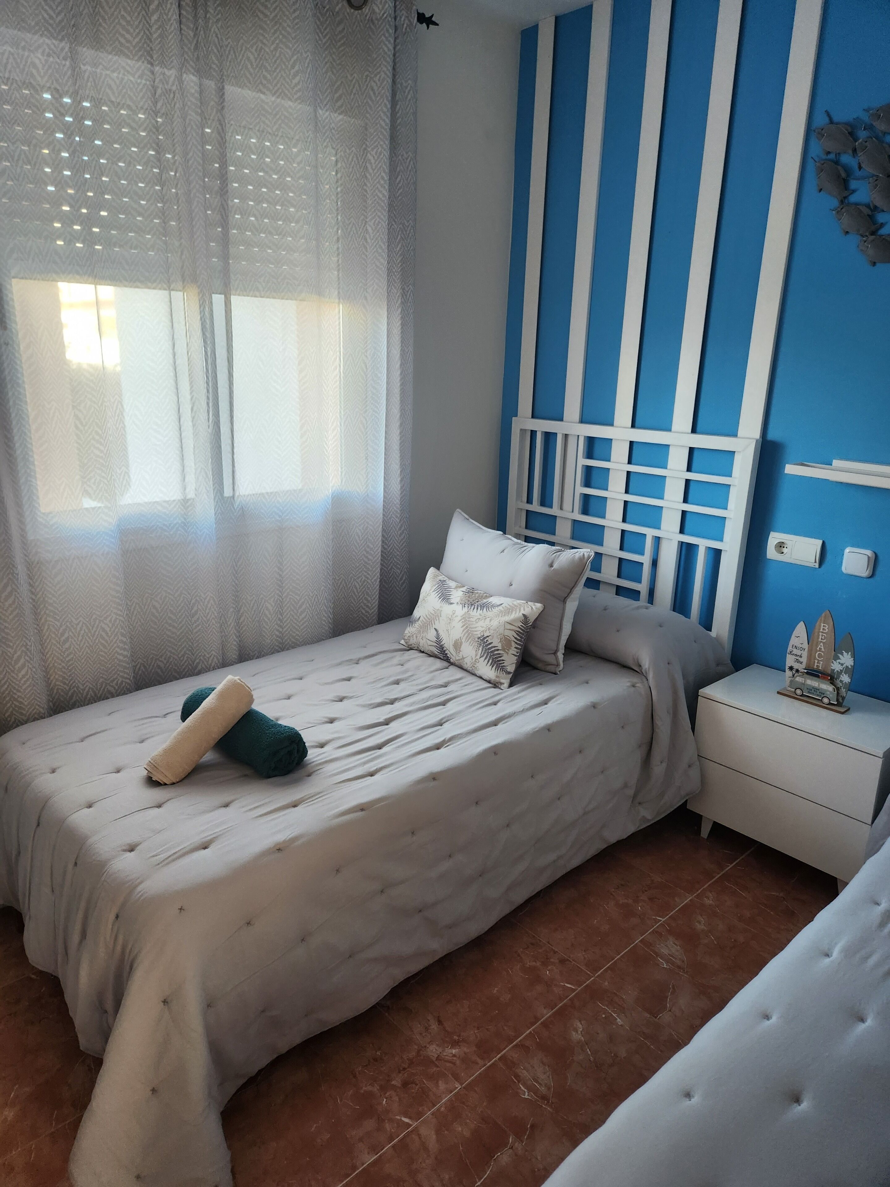 A bedroom with a bed, a nightstand, a window with sheer curtains, and a blue wall with vertical stripes.