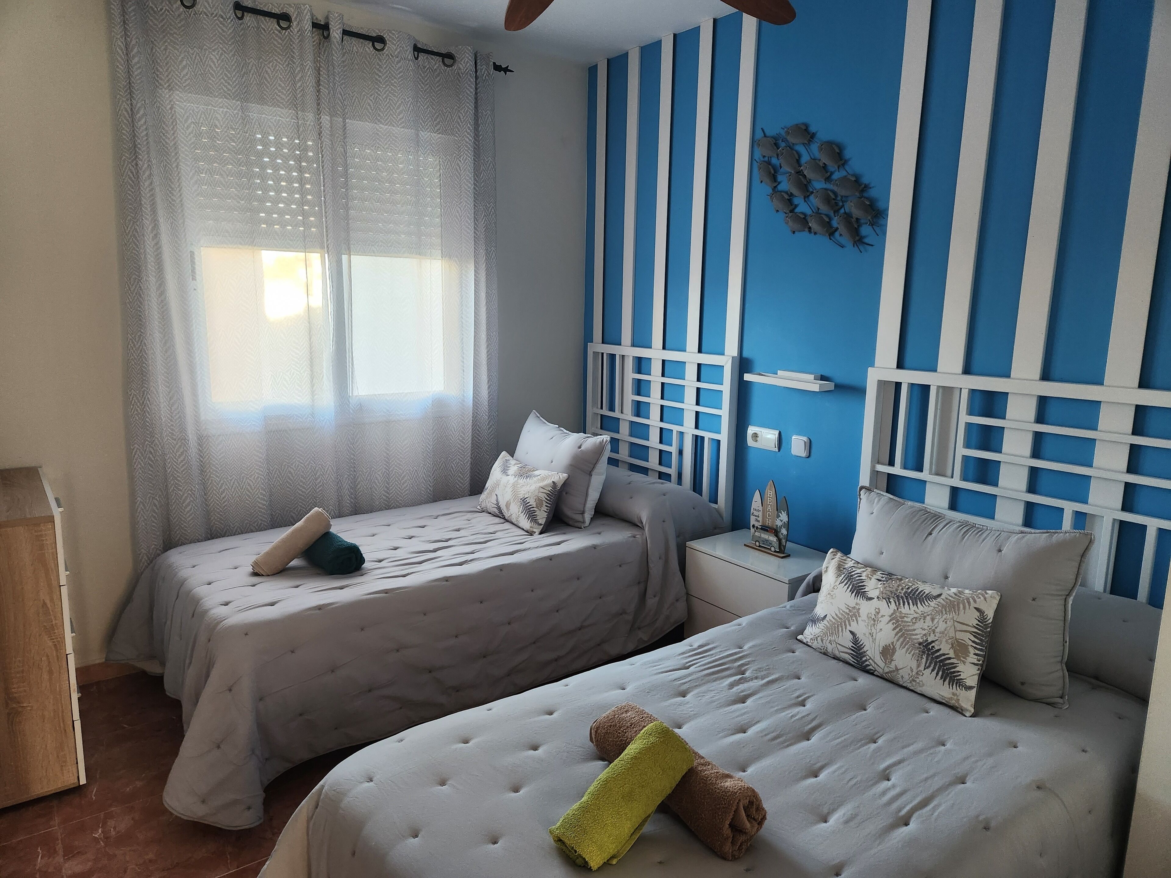 A hotel room with two beds, a window with sheer curtains, a blue and white striped wall, and a small nightstand with a decorative item.