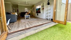Ampla sala de estar com um sofá cinza, uma mesa de centro branca e uma televisão. Piso de madeira e área externa com grama sintética verde.