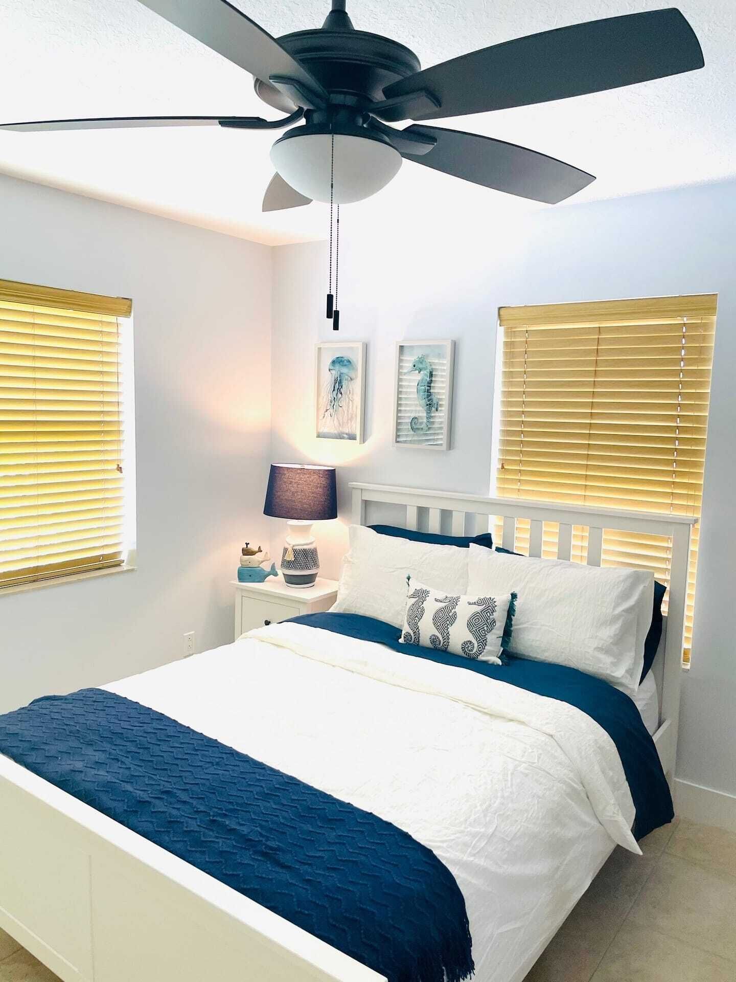 Une chambre à coucher comprenant un lit, un ventilateur de plafond, une table de chevet avec une lampe et deux tableaux accrochés au mur.