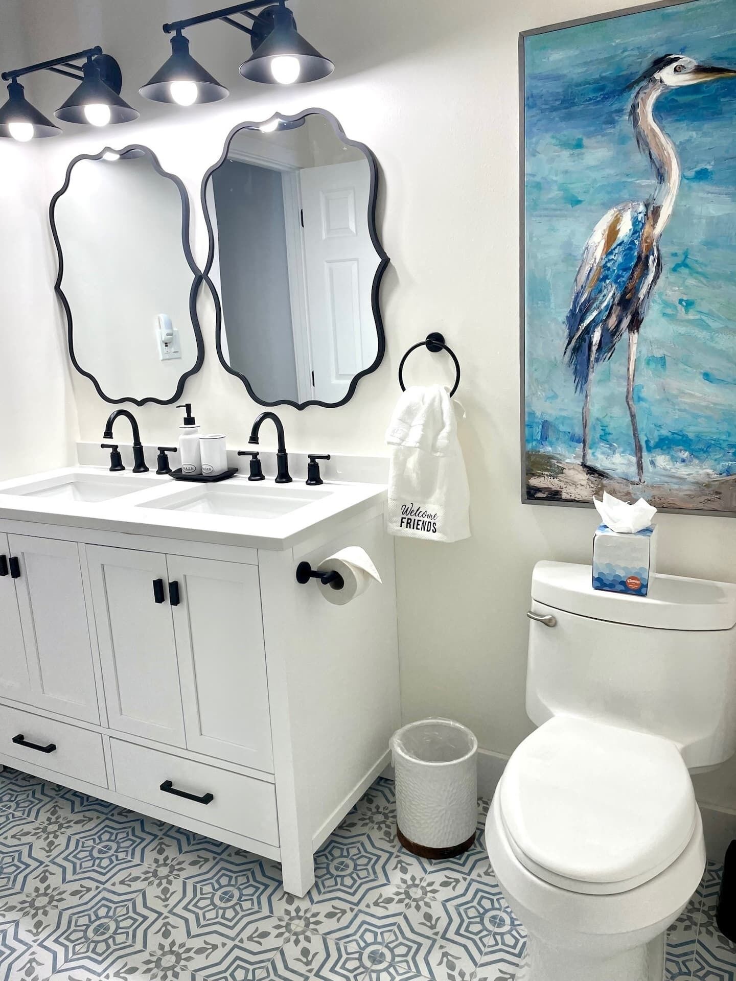 A bathroom with a white vanity, a toilet, a trash can, and a painting of a bird on the wall.