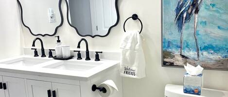 A bathroom with a white vanity, a toilet, a trash can, and a painting of a bird on the wall.