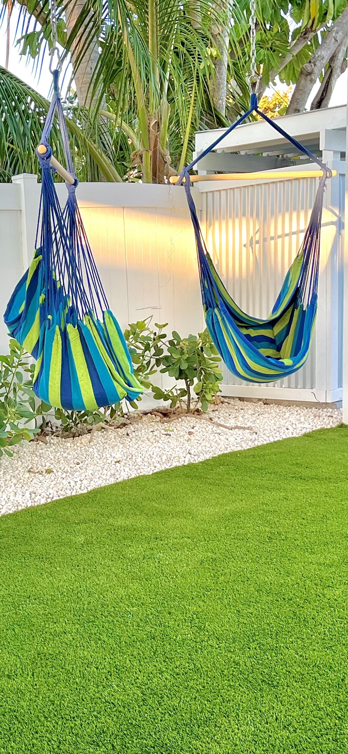 Deux hamacs, l’un bleu et l’autre vert, sont suspendus dans un jardin agrémenté d’une clôture blanche et de plantes vertes.