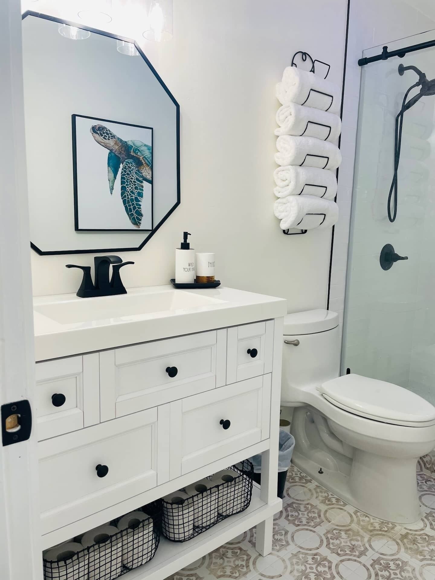 A modern bathroom with a white vanity, a framed sea turtle artwork, a glass shower enclosure, and neatly arranged toiletries.