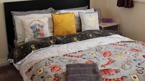 A bed with a Harry Potter themed duvet cover, pillows with various designs, and a bedside table with a lamp and a clock.