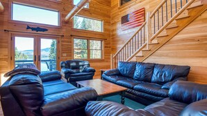 A log cabin interior with a wooden staircase, leather sofas, and a view of the outdoors through large windows. - Dodge Lodge | Private Waterfront on Dodge Pond, Newly Renovated, Near Downtown Rangeley (Rangeley)