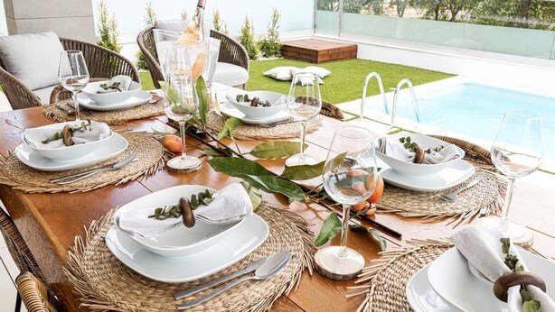 A dining area with a pool view, set tables with white plates, glasses, and wooden utensils, and a wicker chair.